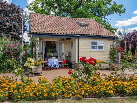 A small cottage with a tiled roof and open double doors leading to a table with two chairs surrounded by flower beds with yellow and red flowers at Norbank Cottage in Bressingham