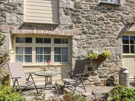A stone wall exterior with two chairs and a table with drinks and flowers a flower basket on the wall and a metal milk container outside at Trevena Crantock near Newquay