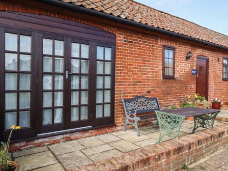 A brick patio with wooden table and metal chairs outside a cottage with large wooden framed windows and wooden door at Barn Owl Cottage Little Glemham near Wickham Market