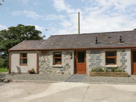 A single-story stone and stucco house with wooden doors and windows and a gravel area in front at Y Beudy Cottage in Clynnog Fawr
