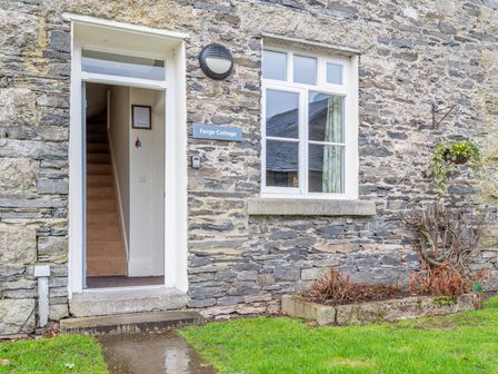 Stone exterior wall of a cottage with an open door showing stairs and a window with a flower basket hanging beside it at Forge Cottage in Graythwaite near Hawkshead