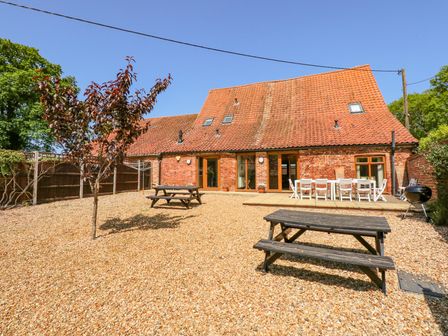 A backyard with a gravel ground a small tree two picnic tables and a brick building with a tiled roof at Hadleigh Farm Barn in King's Lynn