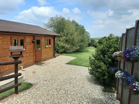A wooden cabin with a gravel path and green lawn surrounded by trees and flowers at Pennylands Willow Lodge in Broadway