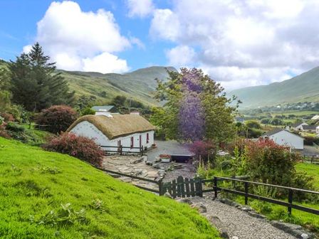 A white thatched cottage with a stone path and green gate surrounded by trees and bushes in a hilly landscape at Granny Kates in Ardara County Donegal