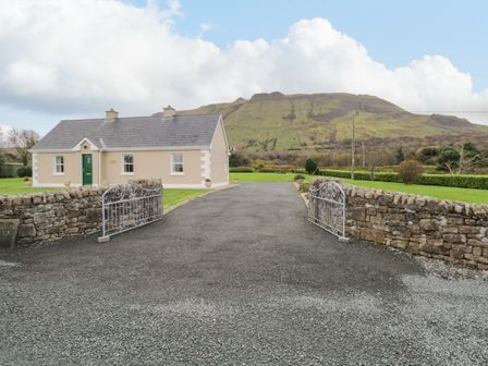 A single story house with a green door and two chimneys with a paved driveway and open metal gates at Tieve Baun Ballintrillick near Kinlough County Sligo