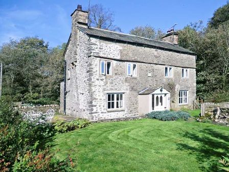 A stone cottage with multiple windows and a green lawn surrounded by trees at Fleshbeck Cottage in Mansergh near Kirkby Lonsdale