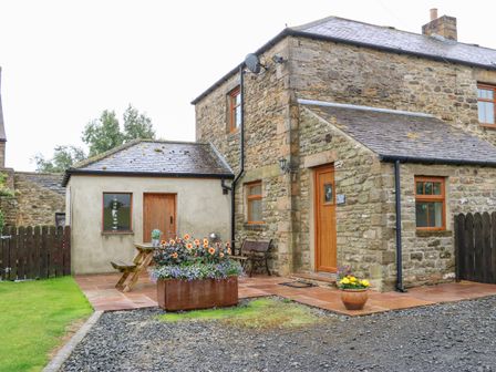 A stone house with wooden doors and windows a bench a picnic table and flower pots outside at The Barn Cottage in Steel near Hexham