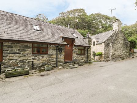 A stone cottage with a slate roof next to a road with trees in the background at Hendoll Cottage 1 Fairbourne near Dolgellau