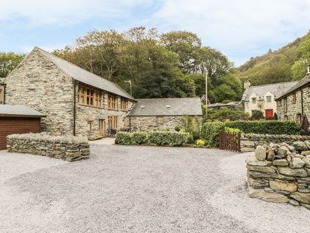 A group of stone buildings with wooden windows and a gravel driveway with stone walls and greenery at Hendoll Barn in Friog near Fairbourne