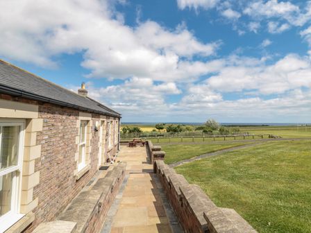 A stone patio with a picnic table beside a brick house overlooking a grassy field and distant horizon at Grey Croft in Embleton