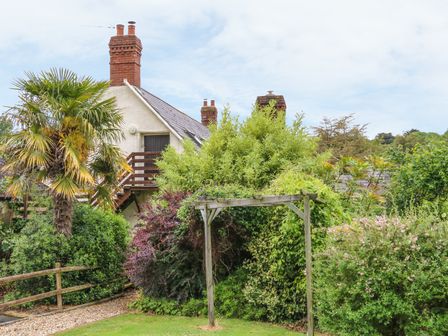 A garden with a wooden pergola surrounded by various bushes and trees with a house and chimneys in the background at Fig Trees - Wibble Farm in Williton near Watchet