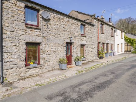 A stone cottage exterior with red framed windows and a door with flower pots on the pavement at Rosemary Cottage in Clawthorpe near Burton-in-Kendal
