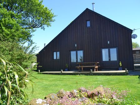 A dark wood barn style house with double glass doors on a lawn with a wooden picnic table and plants at Meader Barn in Peter Tavy near Tavistock