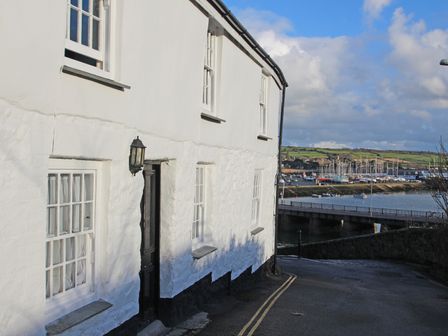 A white building with black door and windows next to a road overlooking a marina with boats and green hills in the background at The Slipway in Penzance