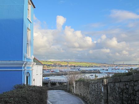 A road with a blue building on the left and a stone wall on the right overlooking a harbor with boats and hills in the background at Mount View in Penzance