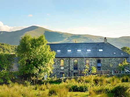 A stone building with arched windows and a slate roof surrounded by greenery and mountains at Capel Dinorwig in Llanberis