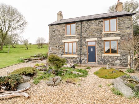 A stone house with wooden framed windows and a blue door surrounded by a garden with gravel paths and grassy areas at Groes Lwyd in Pencoed near Corwen