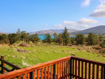 A wooden deck railing overlooking a green field with trees a body of water and hills in the background at Sugarbush in Cahirkeem Cross near Eyeries County Cork