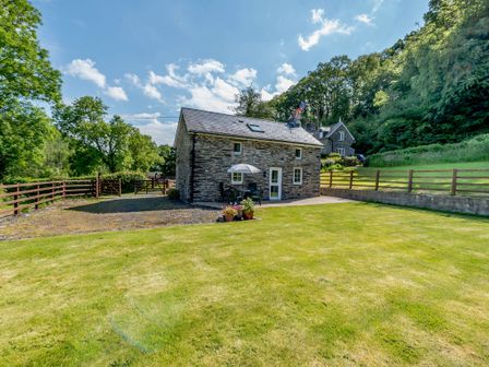 A stone cottage with a patio table umbrella and chairs in a fenced garden with grass and trees at Cwmdu Cottage Cwm-Cou near Newcastle Emlyn