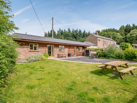 A garden with a wooden picnic table and patio furniture outside a stone and wood building with trees in the background at The Stable @ Rose Cottage in Yorkley
