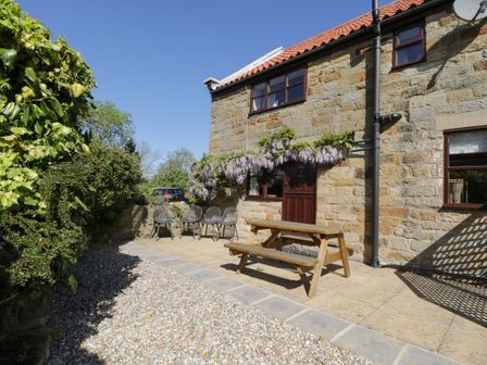 An outdoor patio with a wooden picnic table stone wall and a stone house with purple flowers at Goathland Cottage in Ruswarp near Whitby