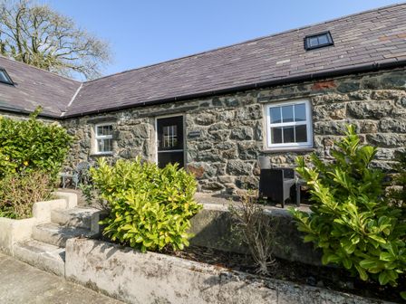 The exterior of a stone cottage with a black door two windows and outdoor seating surrounded by plants at Bwthyn y Dderwen Oak Cottage in Pwllheli