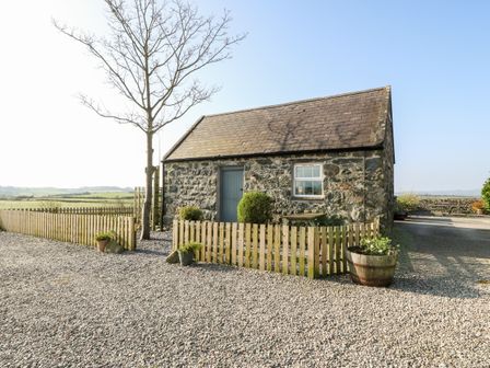 A small stone building with a pitched roof surrounded by a wooden fence and gravel at Bwthyn Celyn in Pwllheli