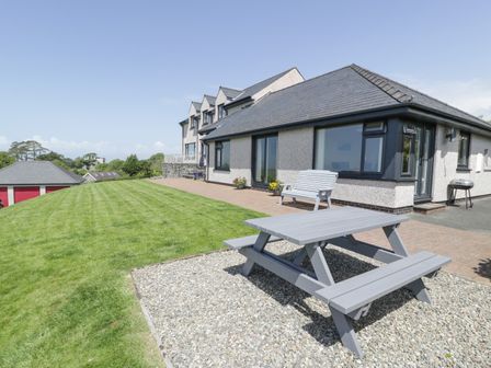 A house with large windows a bench and a picnic table on a gravel area and a grass lawn at Bryn Coch Bach in Dyffryn Ardudwy