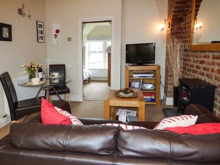 A living room with a brown leather sofa red and white cushions a glass dining table with two chairs a television on a wooden stand and a doorway to a bedroom at Bluebird Loft in Coniston near Hawkshead
