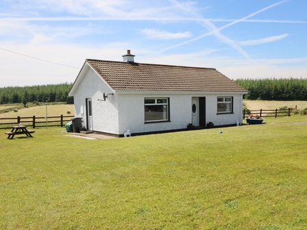 A white single-story house with a tiled roof on a grassy lawn with a wooden picnic table and trees in the background at Clash-Brack in Dungarvan County Waterford