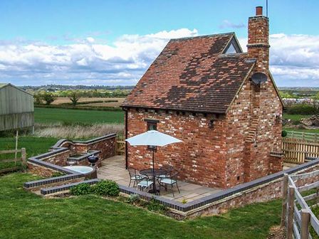 A small red brick house with a tiled roof and chimney surrounded by a low brick wall with a patio table set and umbrella on the patio area at Owl Cottage in Napton-on-the-Hill near Southam