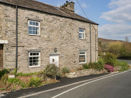 A stone cottage with four windows and a white door next to a road at Duddon Cottage in Broughton-in-Furness