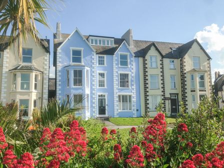 Three connected houses with bay windows and a garden with red flowers in front at Sea View Apartment in Llanfairfechan