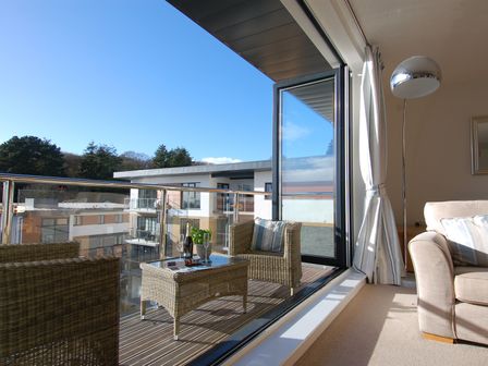 Balcony with wicker chairs and table with glass doors open to a living room with a sofa and floor lamp at 28 Clock Tower Court in Charlestown