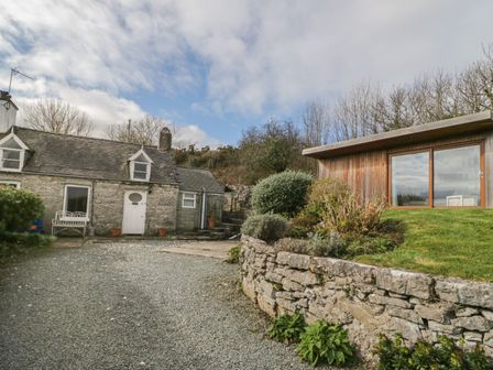 A stone cottage with a white door and windows next to a modern wooden building with large glass doors at Bron Heulog Cottage Llangoed near Beaumaris