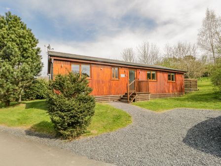 An exterior view of a wooden lodge with steps leading to the entrance surrounded by grass and trees at Cartmel Lodge in Allithwaite near Grange-over-Sands