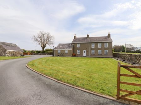 A driveway leading to a two-story house with multiple windows a wooden bench and a stone building with a grassy lawn at Garreg Wen Isaf Bach in Penygroes near Caernarfon
