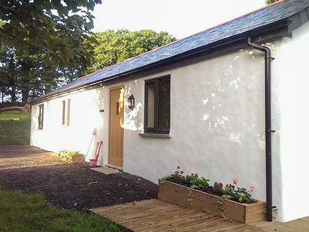 A white single-story building with a wooden door and black window in a garden area at Vine Cottage in St Ewe near Sticker