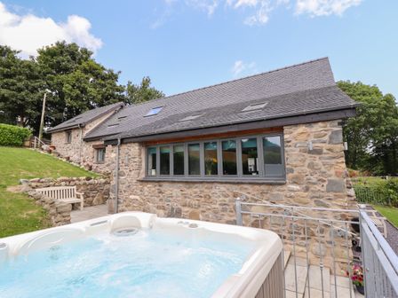 An outdoor hot tub on patio next to a stone house with gray framed windows and a wooden bench nearby at Ysgubor Tyddyn Isaf in Derwen