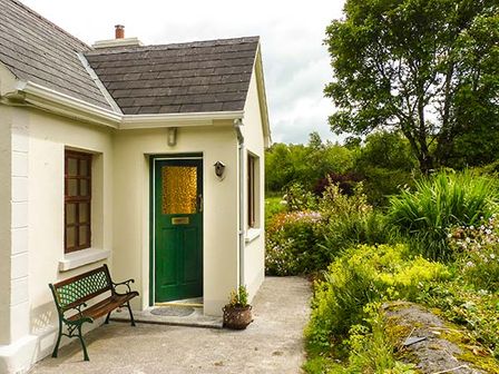 An exterior view of a house entrance with a green door and a bench next to it surrounded by greenery at Hawthorn Farm Cottage in Curry County Sligo