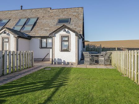 An outdoor patio area with garden chairs and a table beside a white house with wooden fencing at Daisy Cottage in Aldingham