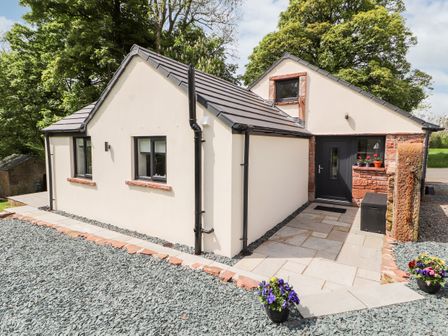 Exterior view of a single-story cream house with black roof and door surrounded by gravel and paved walkway with flowers at Lands Brow Lodge in Wigton