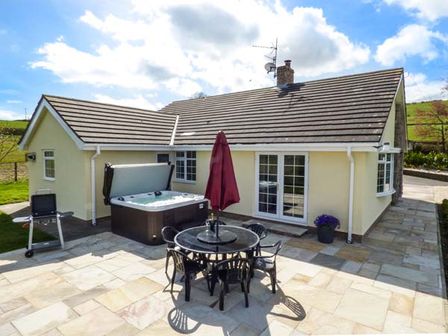 A patio area with a round table and chairs a hot tub a barbecue grill and a yellow house at Bryn Tirion in Betws yn Rhos near Abergele