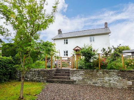 A white house with a tiled roof behind a stone wall garden and wooden gate with trees and gravel path at Gatehouse in Painscastle