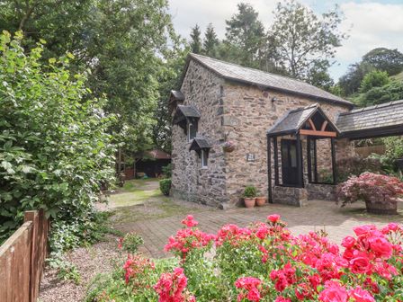 A stone building with a tiled roof next to a paved area and pink flowers at The Old Barn in Llanferres near Ruthin