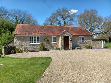 A stone cottage with a red tiled roof a brown door two white windows a bench gravel driveway and green lawn at Magpie Cottage in South Brewham