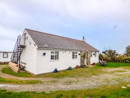 A white cottage with a pitched roof and external staircase on the side with potted plants and chairs on grass at Ty Woods Cottage in Rhoscolyn near Trearddur Bay