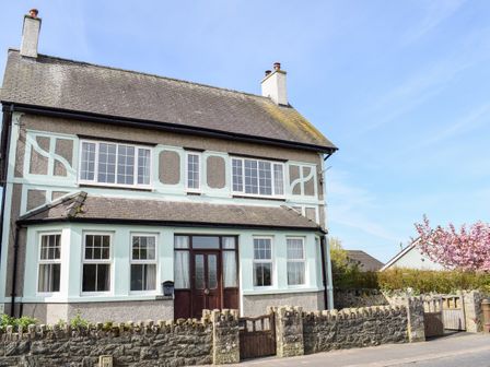 A two-story house with multiple windows a wooden door and a stone wall with a gate in front and a tree with pink blossoms at Minffordd in Newborough