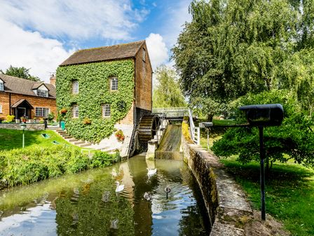 A watermill building covered in ivy beside a river with swans and a water wheel at Grafton Mill in Grafton Flyford near Pershore