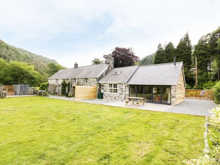 A stone house with a wooden picnic table on a patio and a large lawn surrounded by trees at Hen Gelligemlyn - The Annexe in Ganllwyd